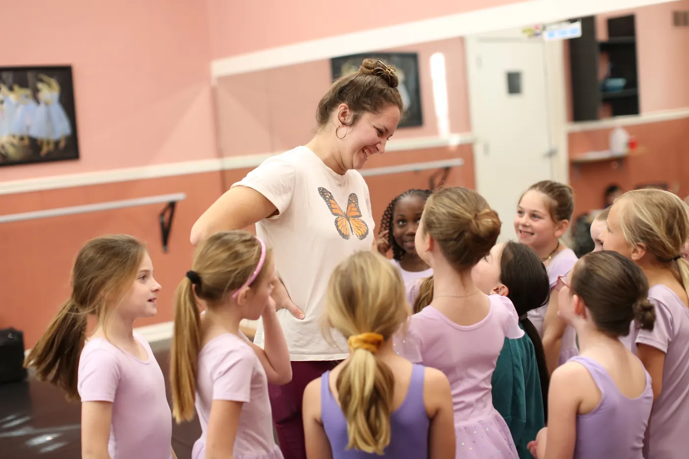 An Elevate instructor smiling as a ring of young dancers in lilac leotards gathers around her in the studio