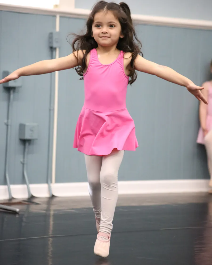 A Creative Movement dancer in a pink leotard balanced on one foot, arms open wide, at the barre in the Elevate studio