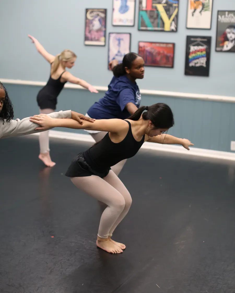 A teen contemporary dancer in a deep hinge, arms swept behind, classmates mirroring the shape