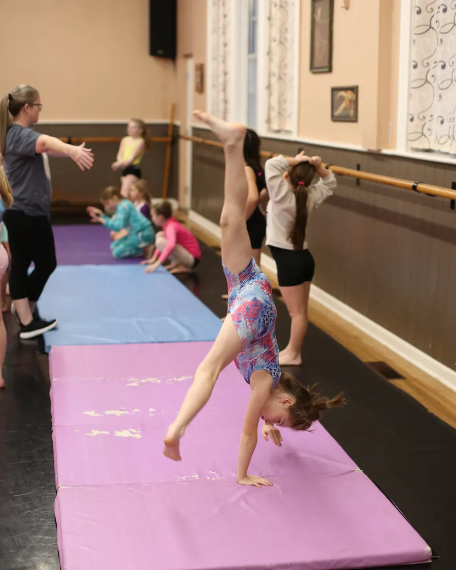 A young Elevate tumbler upside down in a cartwheel on the pink mat, legs straight overhead, classmates waiting along the mats behind her