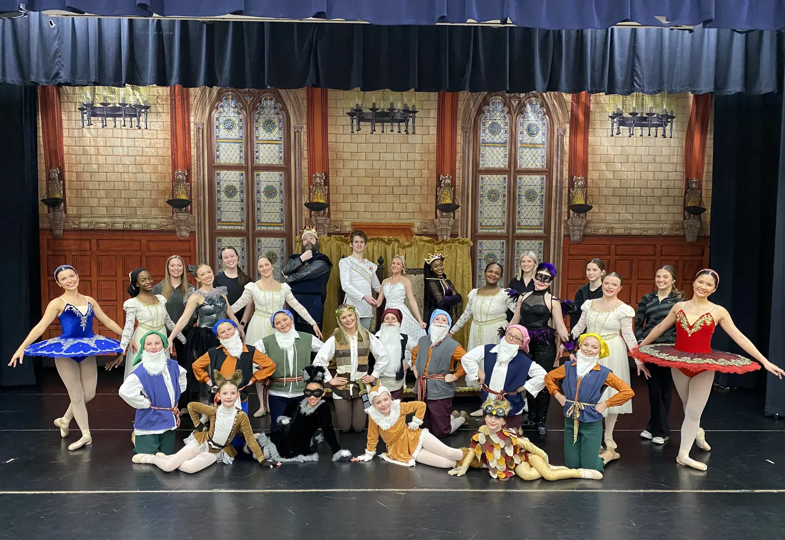 The full cast of a Dance Theatre South ballet posed on stage in front of a painted castle hall, with dancers in blue and red tutus at either end and the youngest dancers kneeling in front in storybook costumes