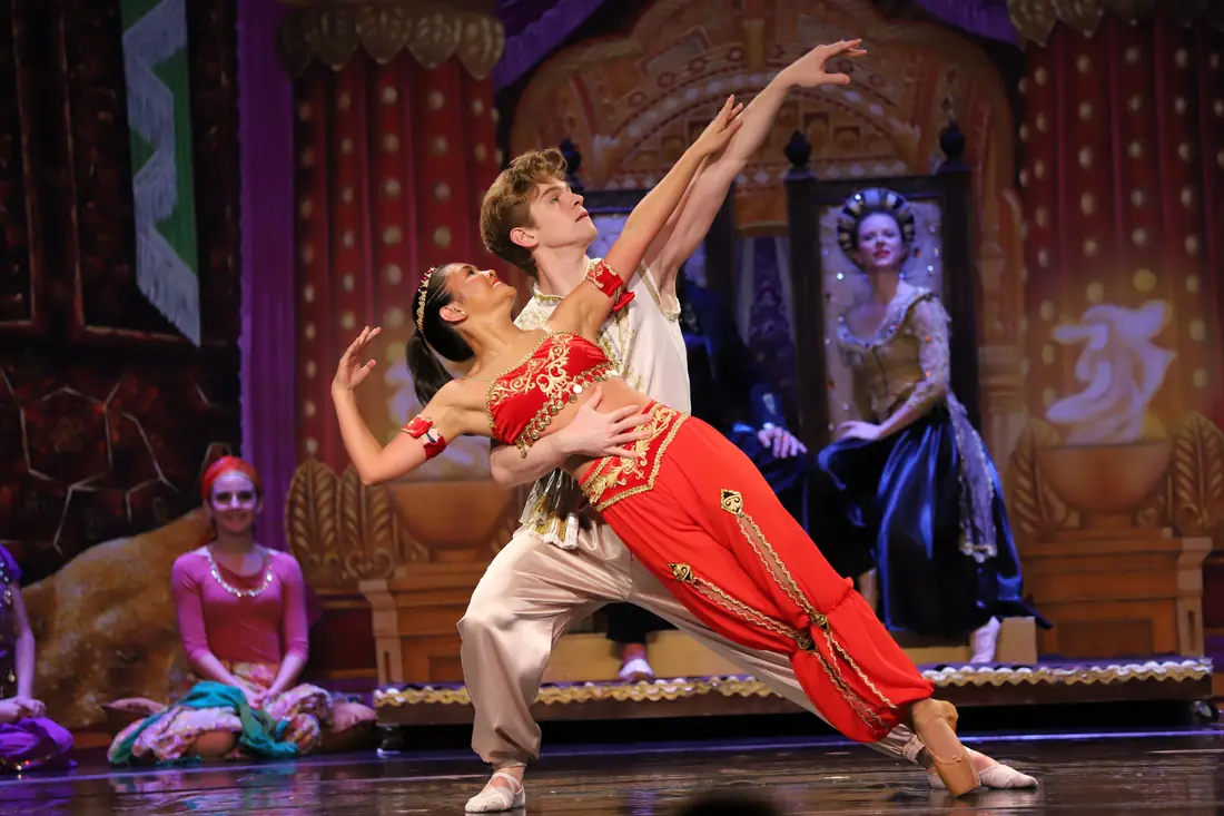 A dancer in red arching back into her partner's arms in a deep lunge while the royal court watches from behind