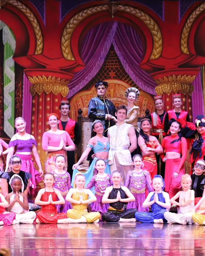 The full cast of a Dance Theatre South ballet posed on stage in jewel-toned costumes, the youngest dancers seated in front
