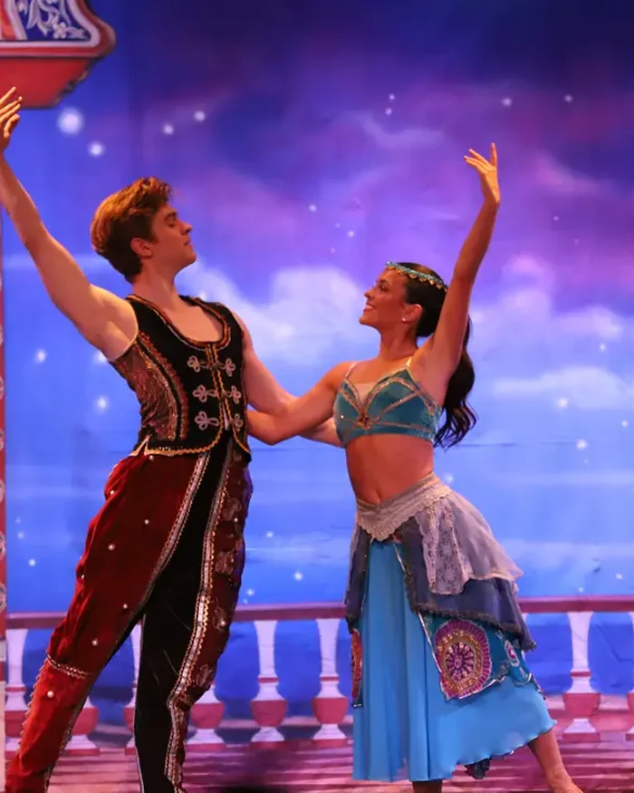 Two lead dancers in jewelled costumes reaching their arms overhead together against a starry night backdrop