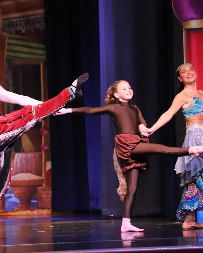 A dancer kicking one leg high as two dancers in flowing skirts follow him across the stage hand in hand