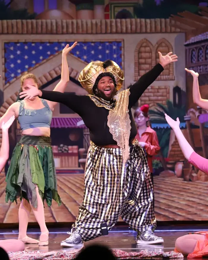 A dancer in a gold turban throwing his arms wide among an ensemble in jewel-toned skirts