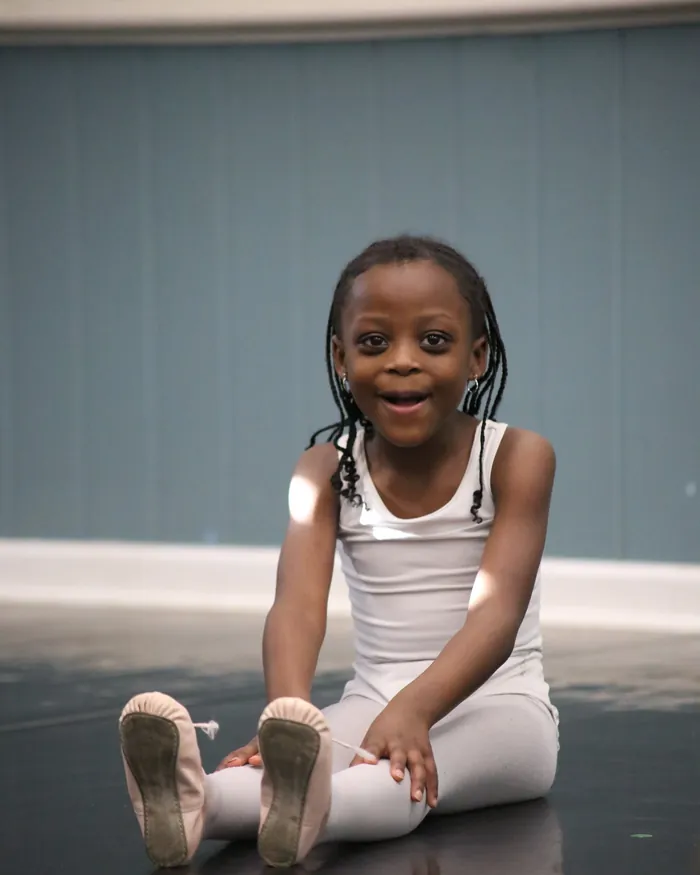 A beaming young dancer in a white leotard sitting on the studio floor with her legs out in front of her