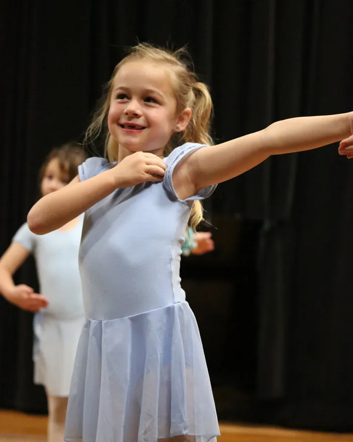A smiling young dancer in a periwinkle leotard and skirt stretching one arm out while her classmate dances behind her