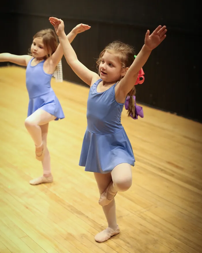 Two young dancers in periwinkle leotards balancing on one leg with their arms raised