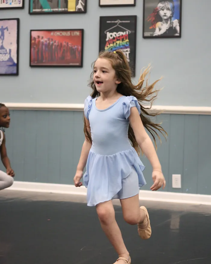 A young dancer in a periwinkle leotard skipping across the studio, her hair flying behind her