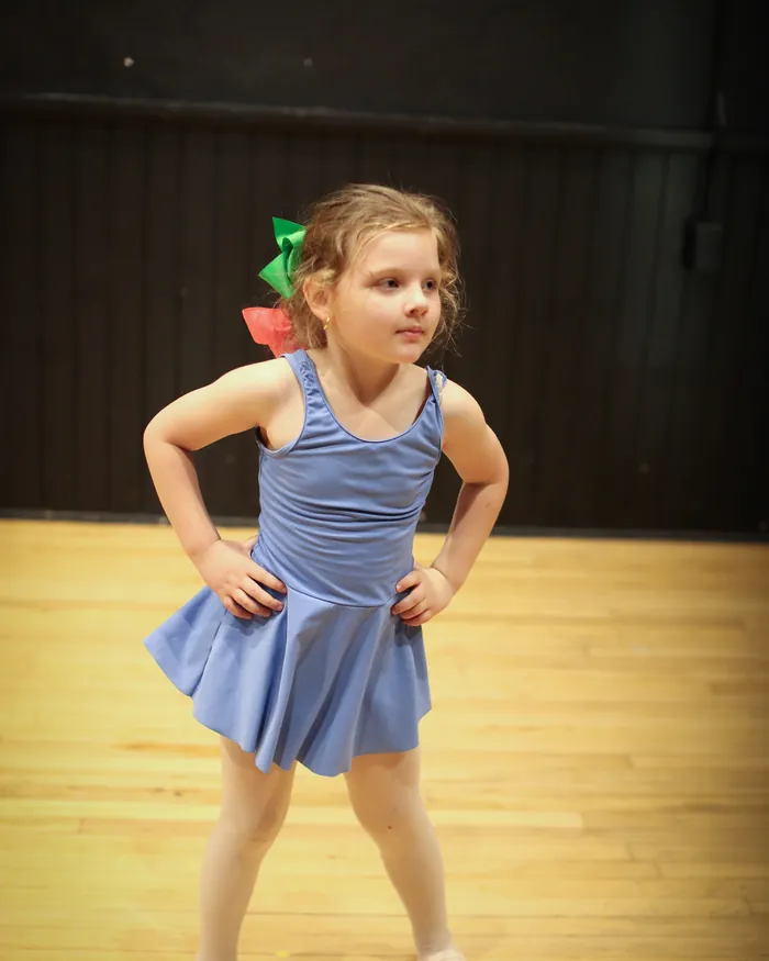 A young dancer with a green hair bow standing with her hands on her hips on the studio floor