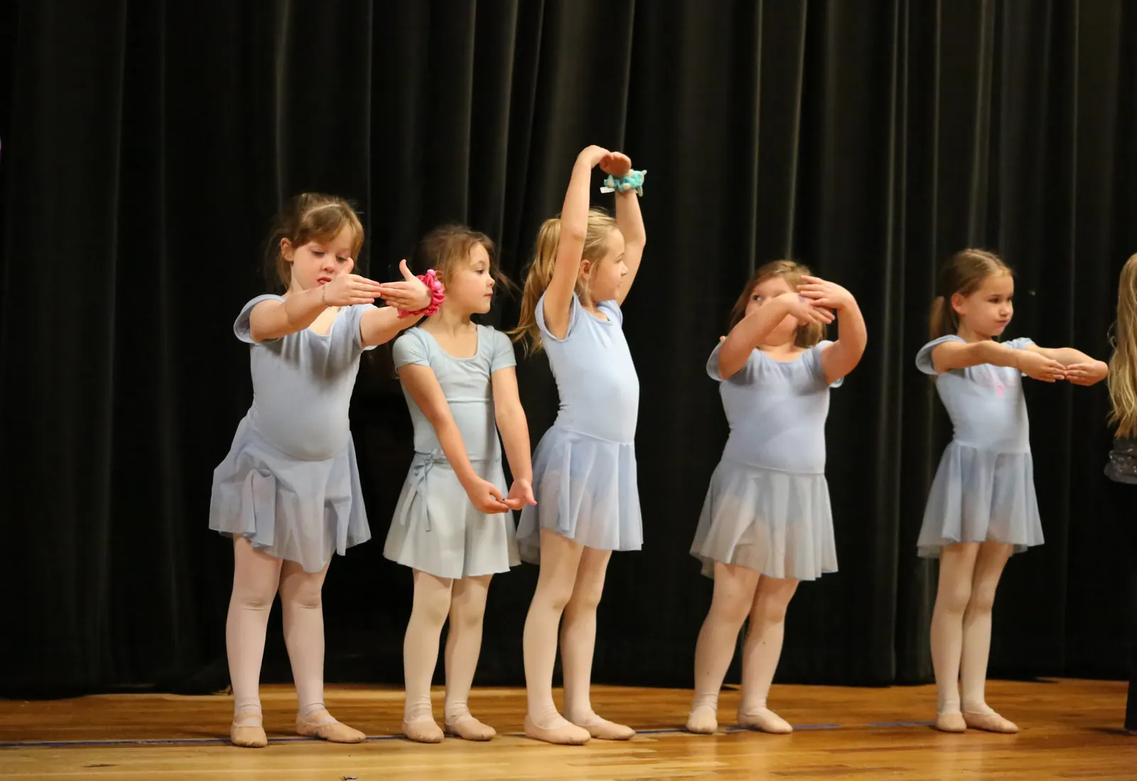 A line of Pre-Ballet dancers in periwinkle leotards and skirts practicing arm positions together on the studio stage