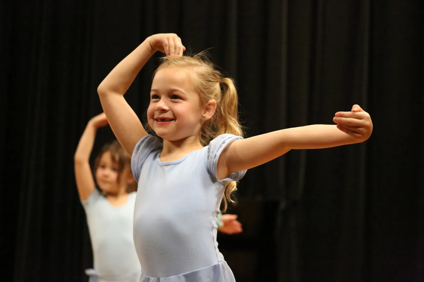 A smiling Pre-Ballet dancer in a periwinkle leotard holding one arm curved overhead and the other out to the side
