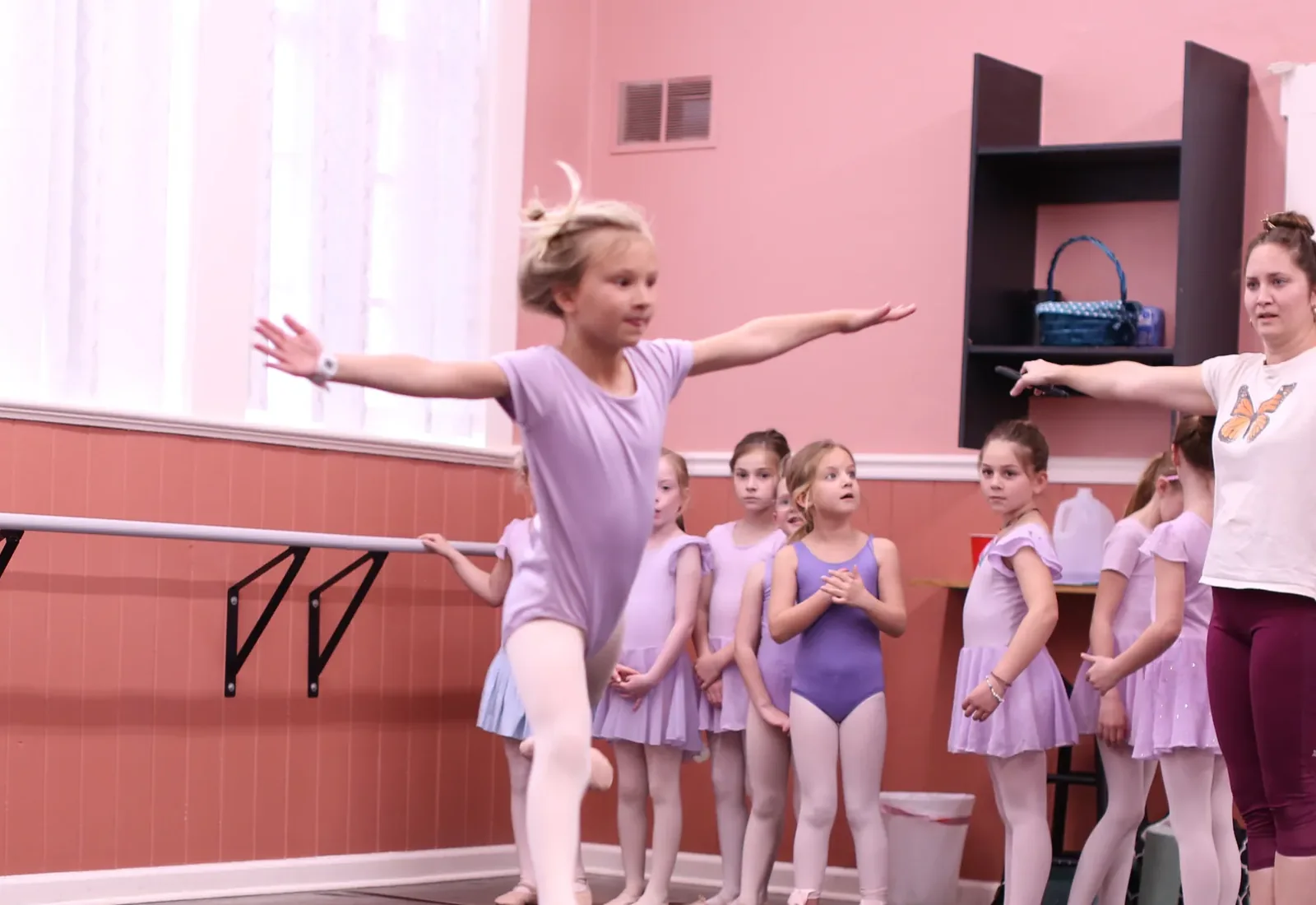 A young Elevate dancer in a lilac leotard extending both arms while her classmates watch from the barre