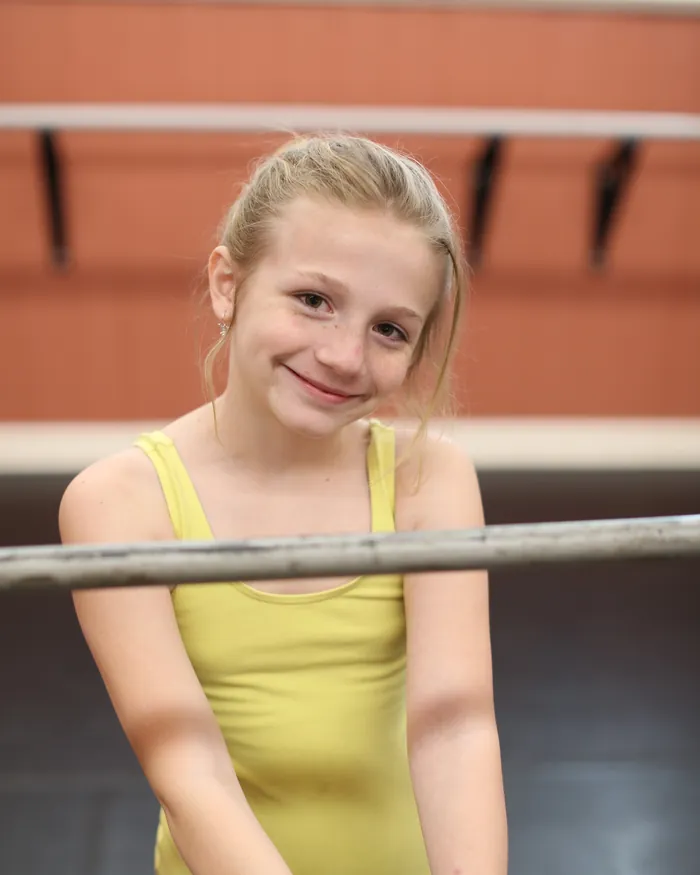 A dancer in a lemon leotard smiling as she leans on the ballet barre