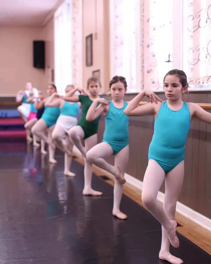 A line of ballet dancers in teal leotards at the barre, one leg lifted in a passe