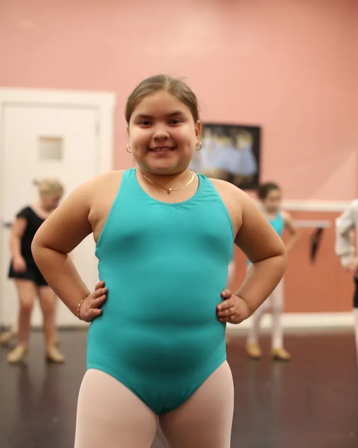A smiling dancer in a teal leotard standing with her hands on her hips