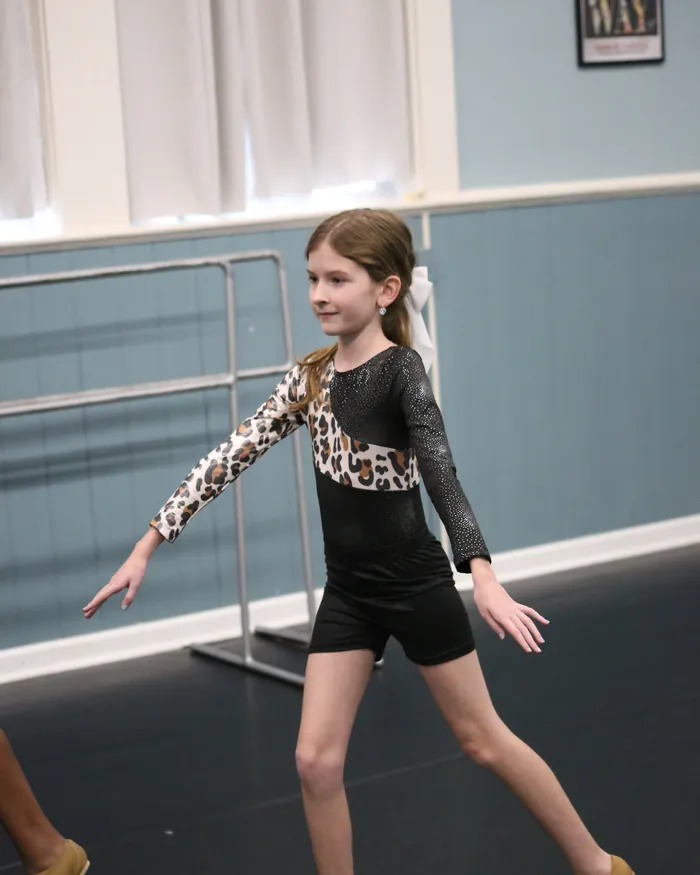 A dancer in a leopard-print leotard smiling as she steps out from the barre with one arm reaching forward