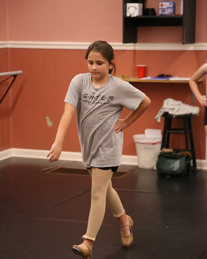 A dancer in tan tap shoes stepping through a tap combination with one hand resting on her hip