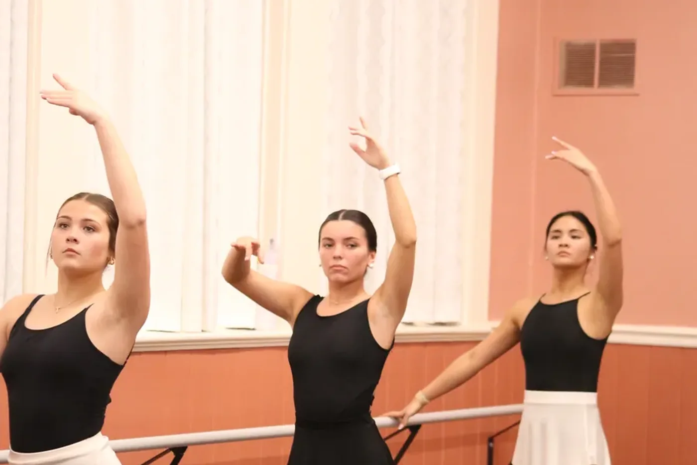 Three teen dancers in black leotards holding a position at the barre with one arm raised overhead
