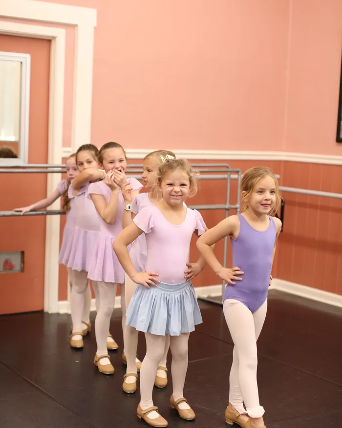 Four young dancers in pink and lilac leotards standing together at the barre, hands on their hips