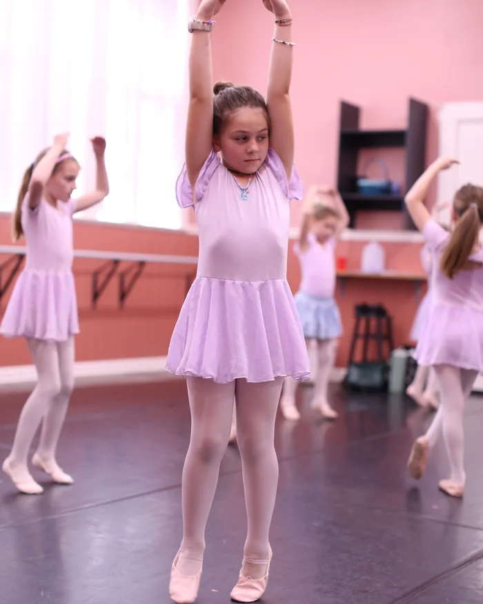 A young dancer in a pink leotard holding both arms curved above her head in the studio
