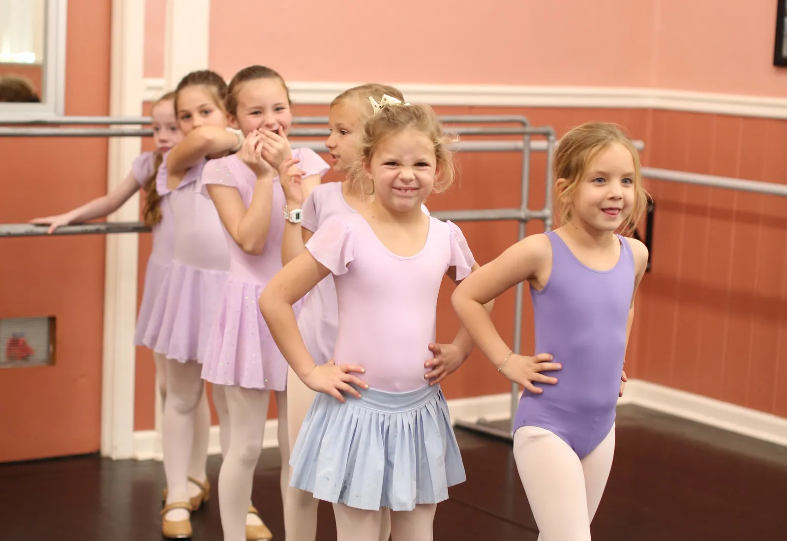 A line of young Elevate dancers in lilac and purple leotards standing at the barre with their hands on their hips, smiling toward the front of the studio