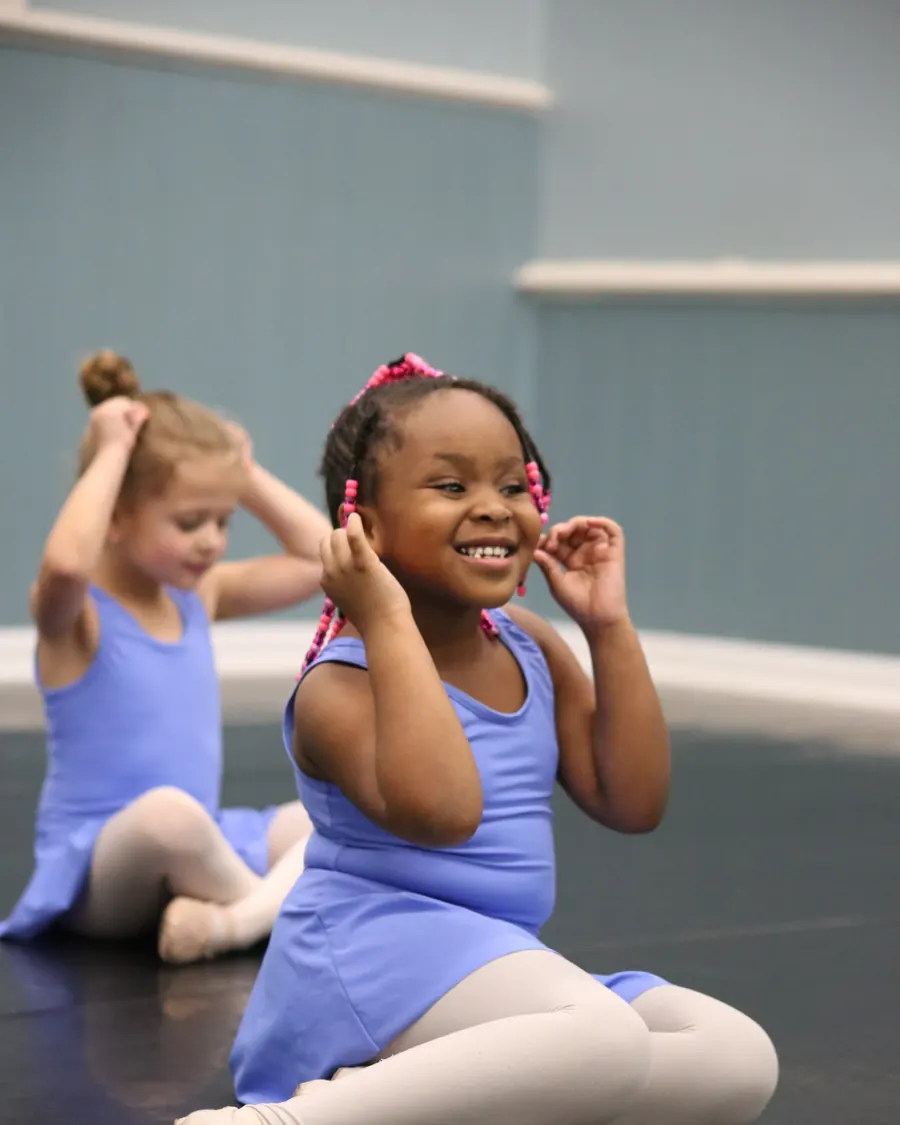 Two young Elevate dancers sitting together on the studio floor, the nearest one laughing as she tucks her hair back
