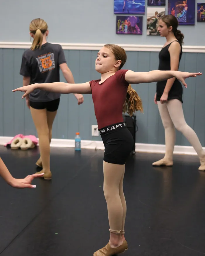 A jazz dancer in a maroon leotard stepping forward with her arms stretched wide