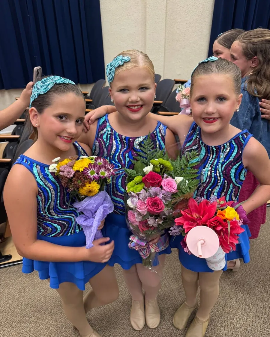 Three young dancers in blue sequined costumes holding flower bouquets in the audience after the show