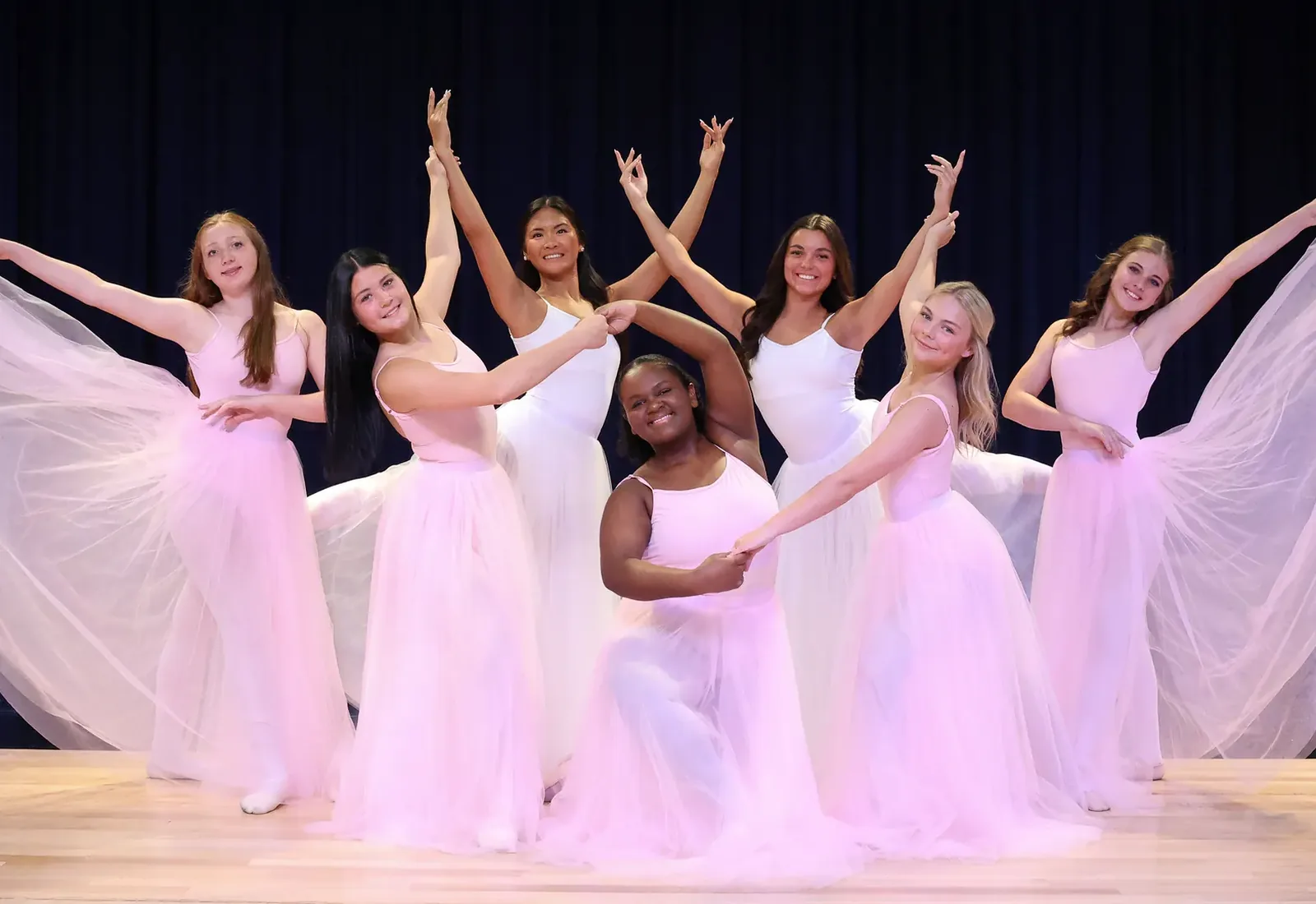 Elevate dancers in pale pink and white costumes posed together on the showcase stage with their arms raised