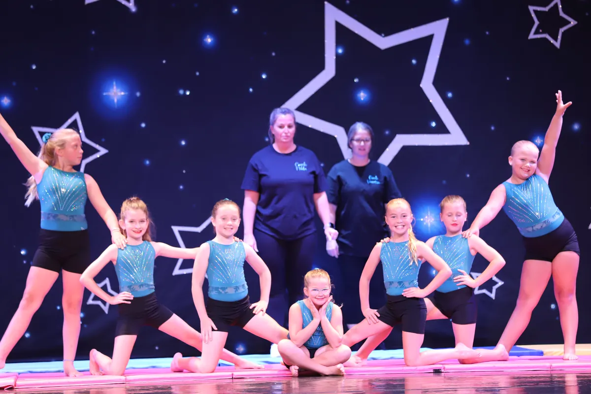 An Elevate tumbling group in teal costumes posed together on the showcase stage with both of their instructors behind them