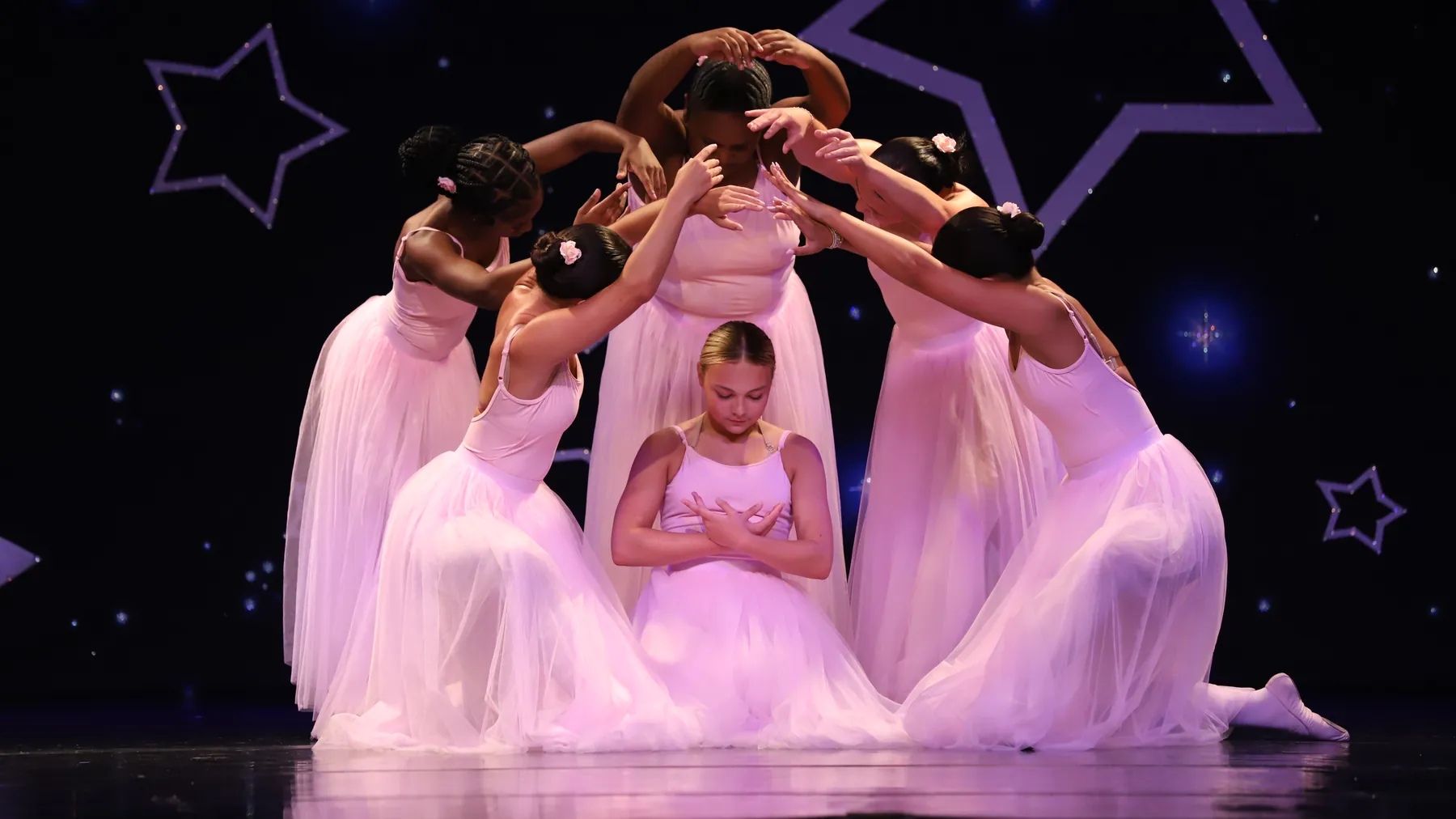 Elevate ballet dancers in long pink romantic tutus gathered around a kneeling soloist under stage lights