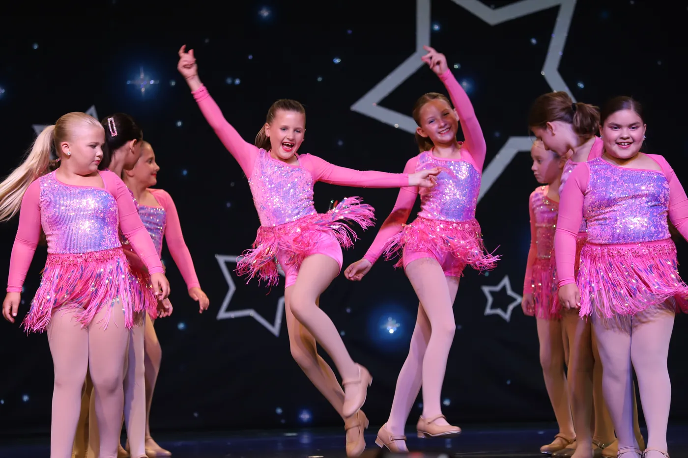 Elevate dancers in pink fringed costumes mid routine, grinning, under a starry showcase backdrop
