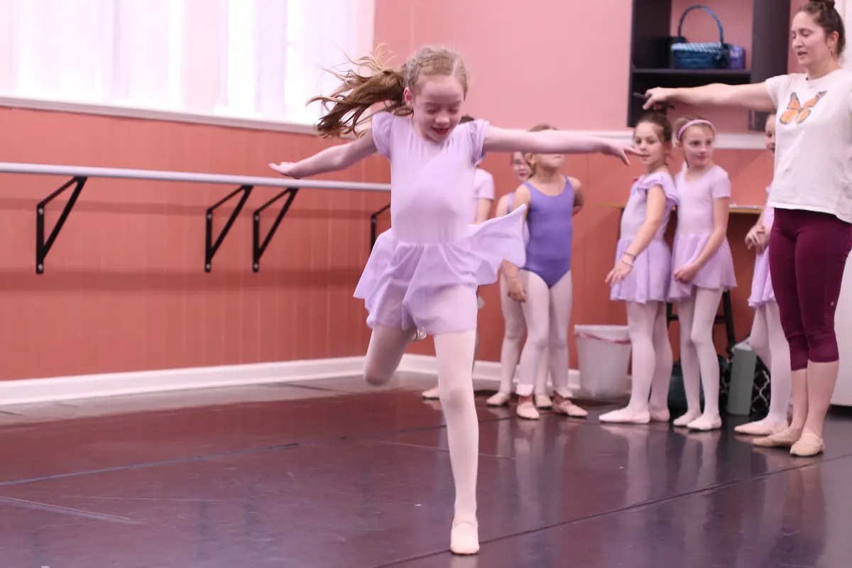 A young Elevate dancer leaping across the studio floor with arms outstretched while classmates and an instructor watch from the barre