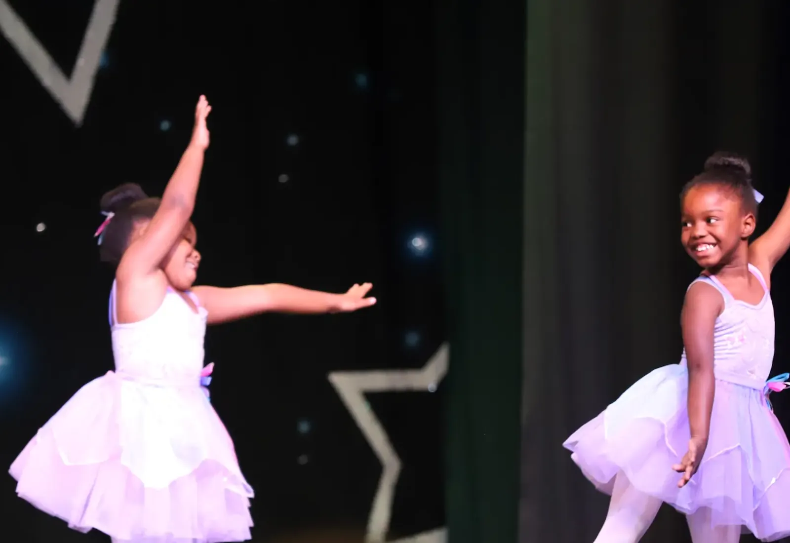 Two of Elevate’s youngest dancers in pale tutus on the Spring Showcase stage, one reaching an arm overhead and the other turning to grin at her