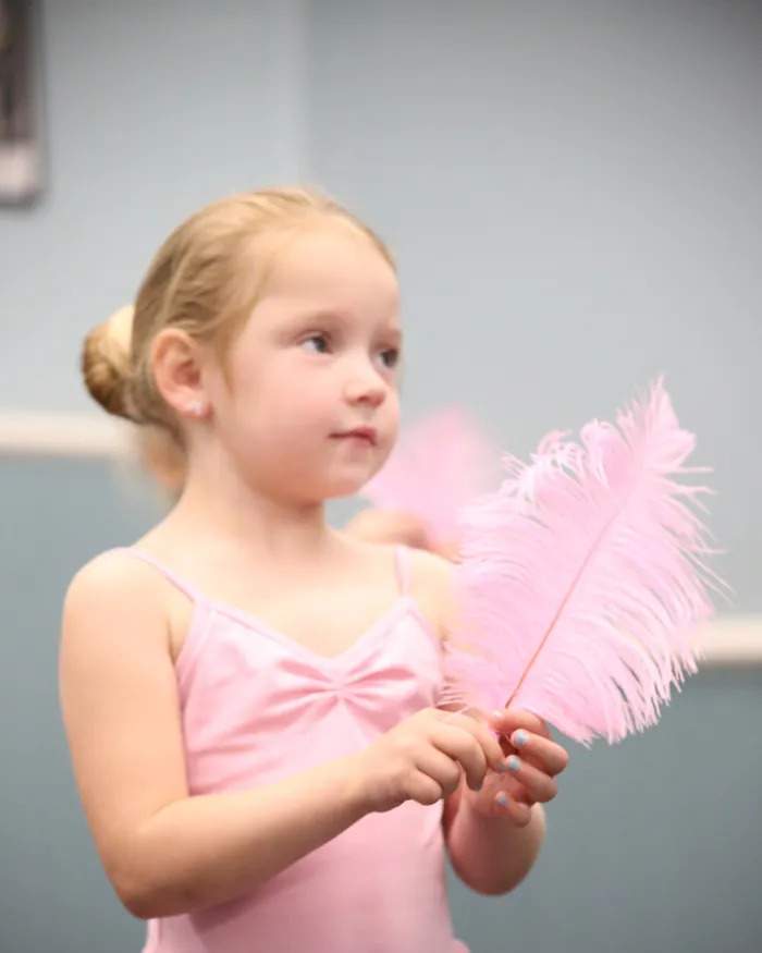 A young dancer in a pink leotard holding a pink feather and watching her teacher