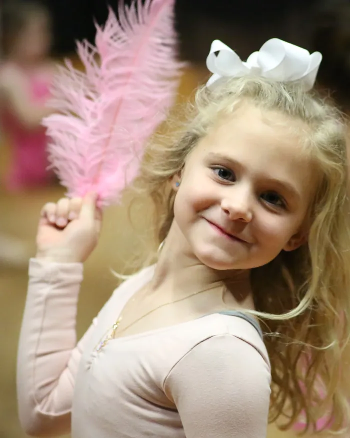 A smiling young dancer with a white hair bow holding a pink feather up beside her face
