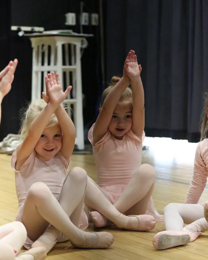 Two young dancers in pale pink leotards sitting cross-legged with both arms raised overhead