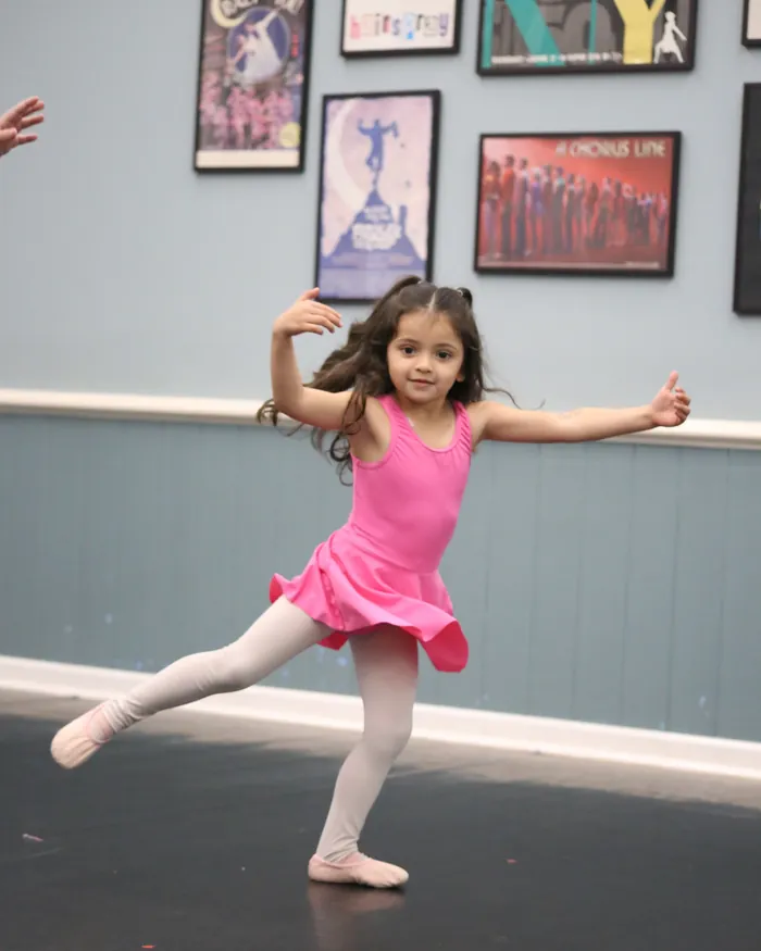 A young dancer in a pink dress leaping across the studio floor with her arms open wide