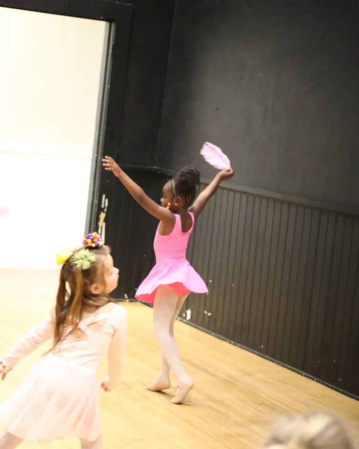 Young dancers moving across the studio with pink feathers, one reaching high toward the wall
