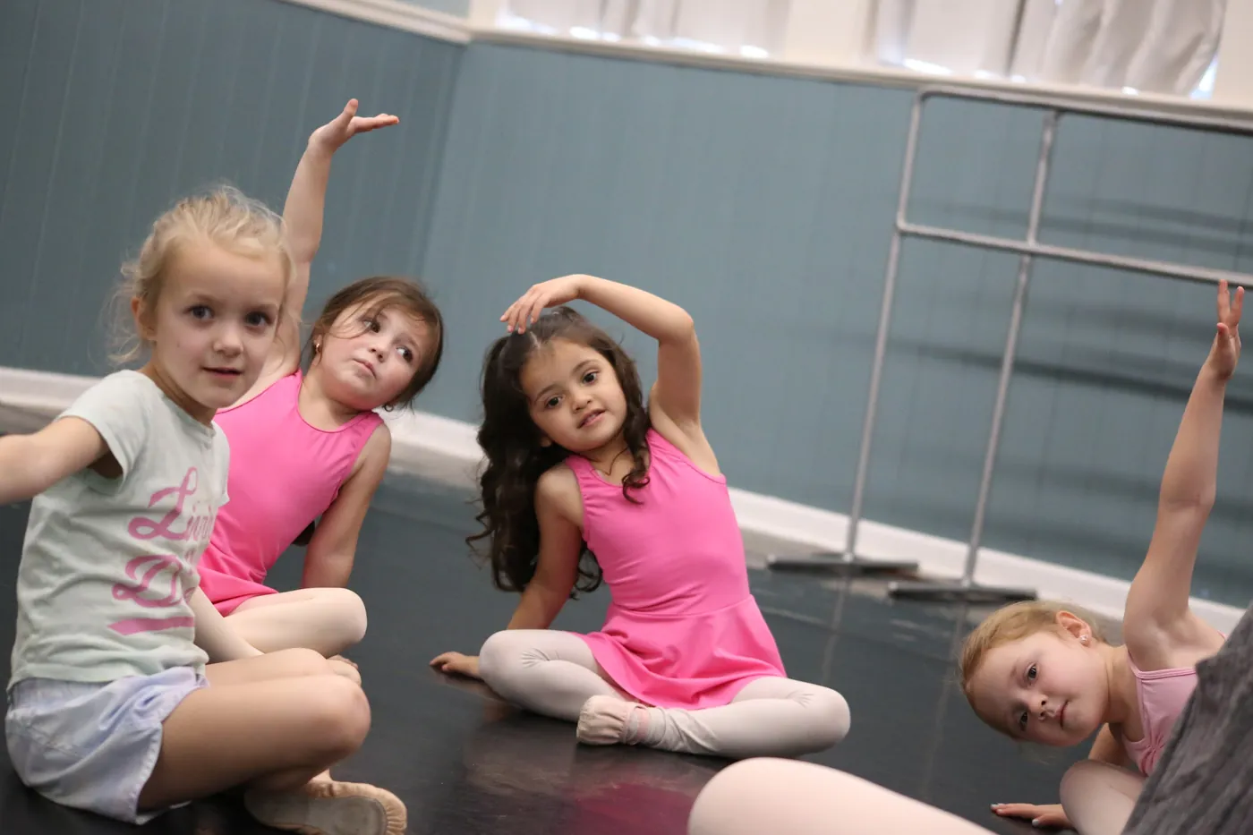 Young dancers sitting on the studio floor stretching one arm up and over, smiling toward the camera